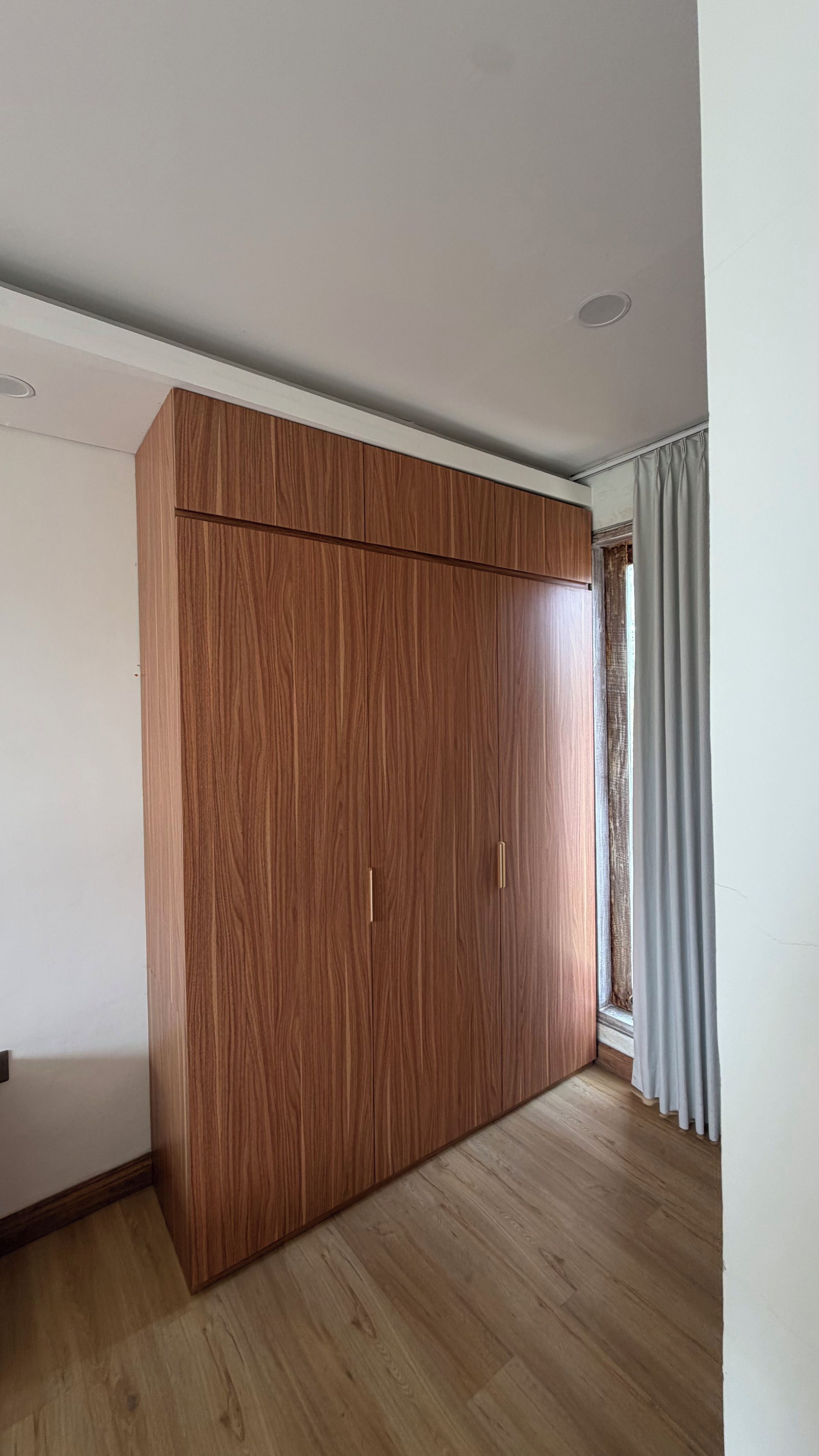 A room with a large wooden wardrobe, a window with curtains, and a wooden floor.