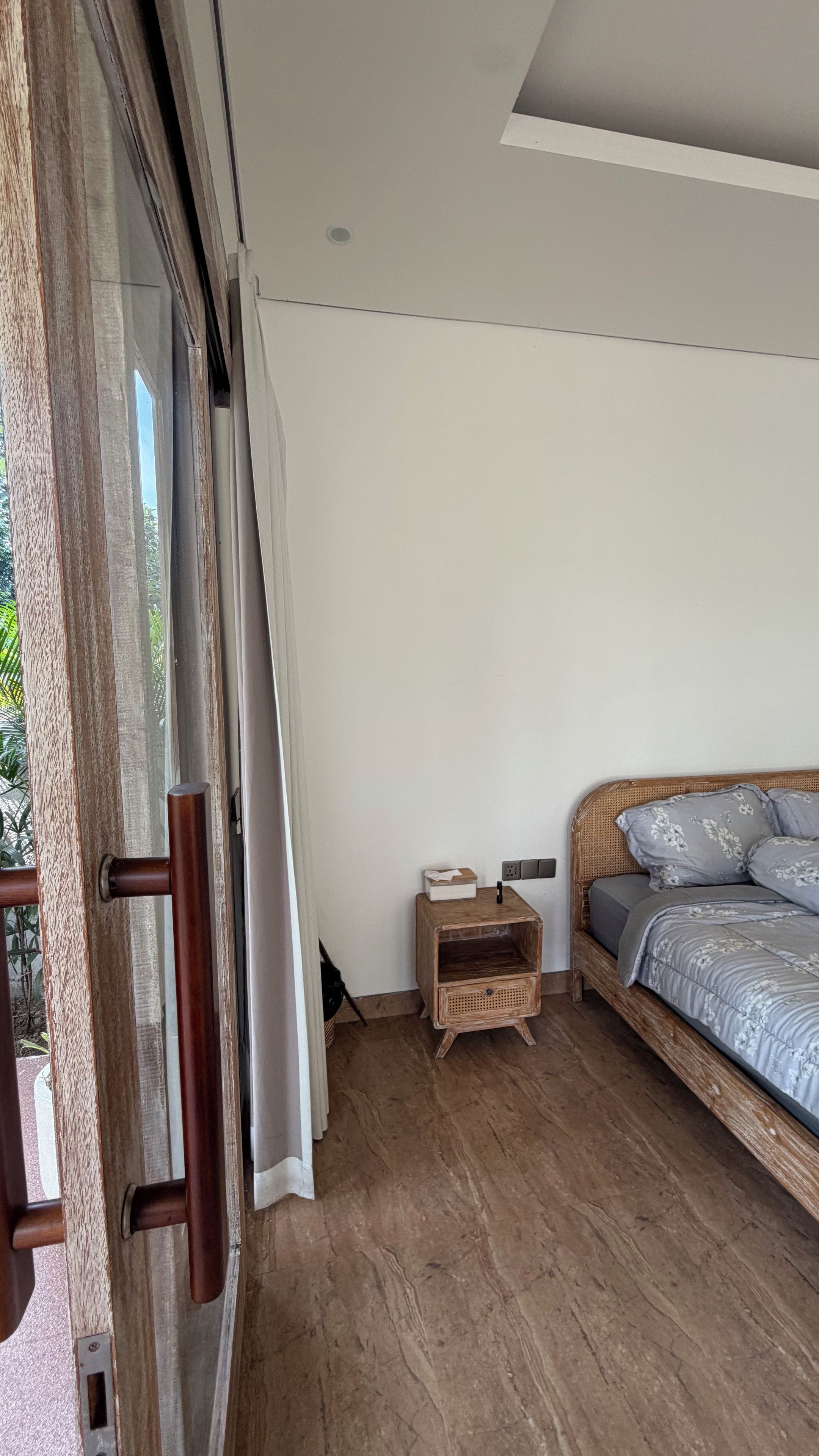 Une chambre avec un plancher en bois, un lit recouvert d’une literie à motifs floraux, une table de chevet et une porte-fenêtre coulissante donnant sur un espace extérieur.