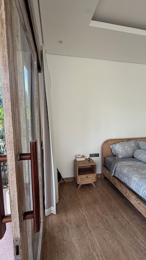 Un dormitorio con piso de madera, cama con ropa de cama floral, mesita de noche y puerta corrediza de vidrio que da a un espacio exterior.