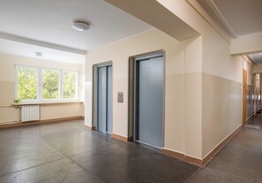 A hallway with two elevators, a radiator, and a window with a view of trees. - Wola Golden Harmony Studio (Warszawa)