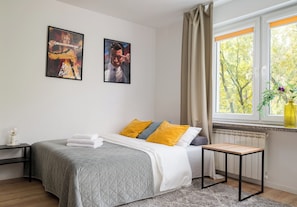 A bedroom with a bed, a bedside table, a window with curtains, and two wall-mounted pictures. - Wola Golden Harmony Studio (Warszawa)