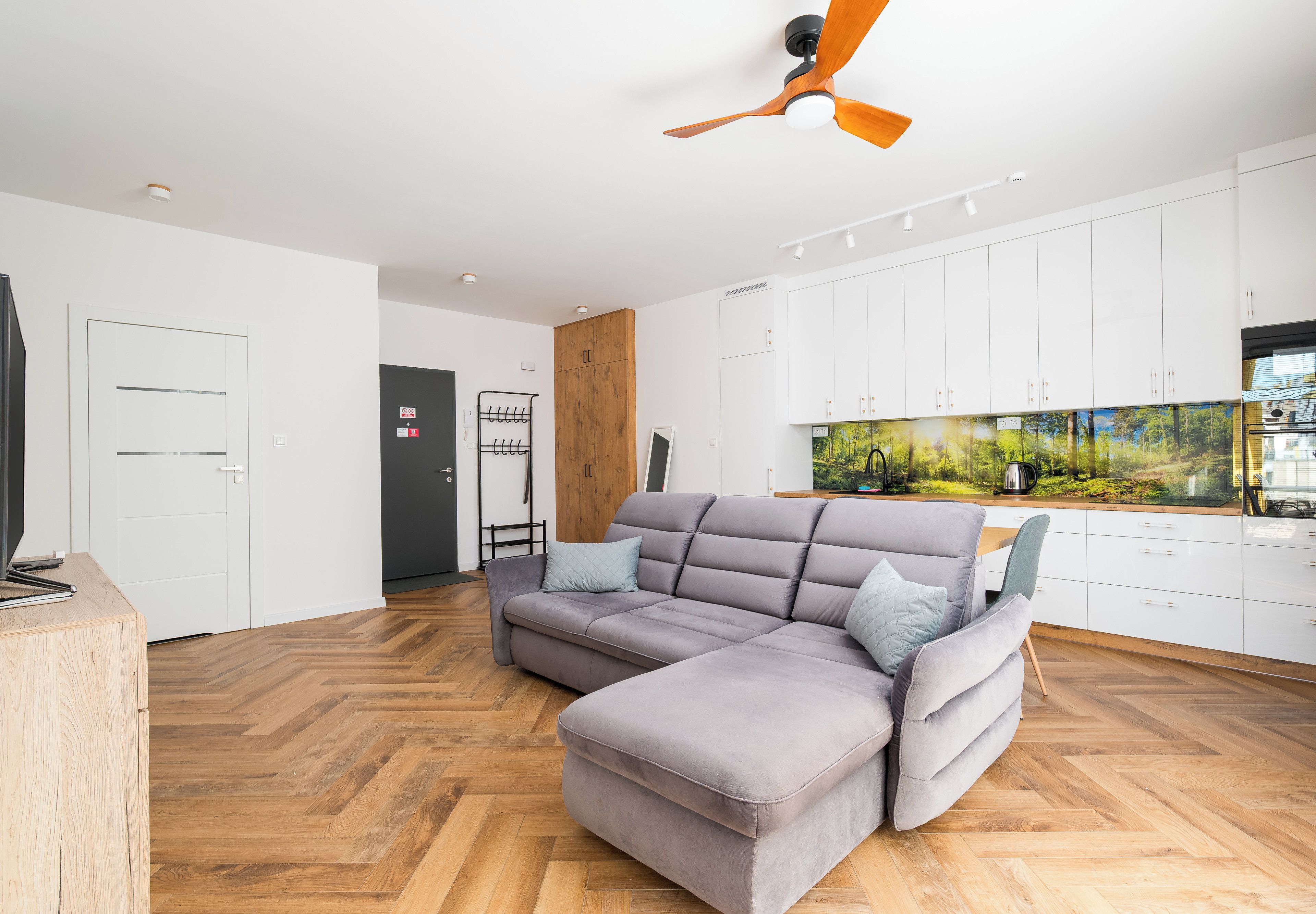 Ein modernes Wohnzimmer mit einem grauen Ecksofa, Holzboden im Fischgrätmuster, weißen Schränken und einem Deckenventilator.