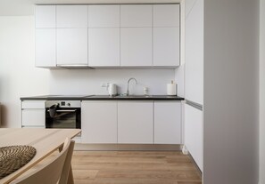 A compact kitchen with white cabinets, a stainless steel oven, a black countertop, and a wooden dining table. - Browary Warszawskie Velvet Studio (Warszawa)