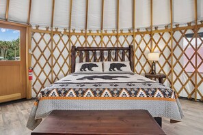 A room with a bed, a wooden headboard, a nightstand with a lamp, and a window with a view of palm trees.