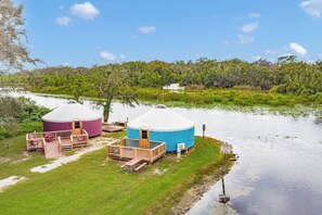 Two yurts with wooden decks and a small dock, situated by a calm body of water with a forested area in the background.
