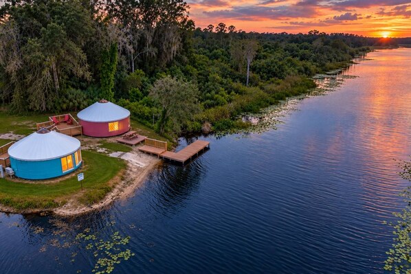 Two yurts by a calm river with a sunset in the background.