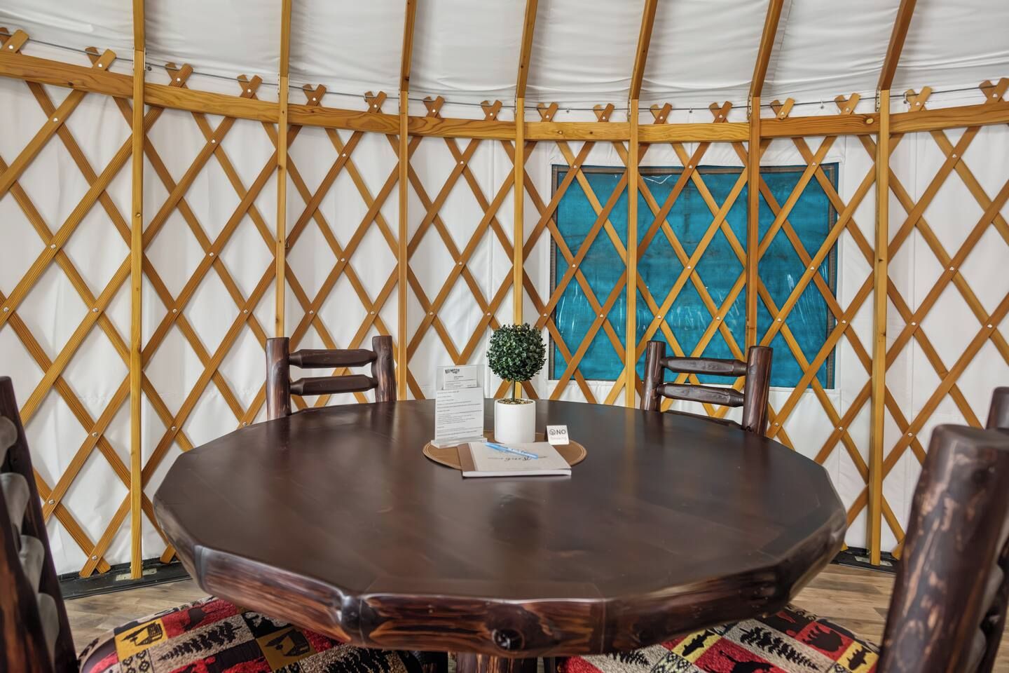 Een ronde houten tafel met stoelen in een tentvormige constructie met een lattenwand en een blauw raam.