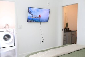 Ett rum med en säng, en tvättmaskin, en tv som är monterad på väggen och ett badrum som syns genom en öppen dörr.