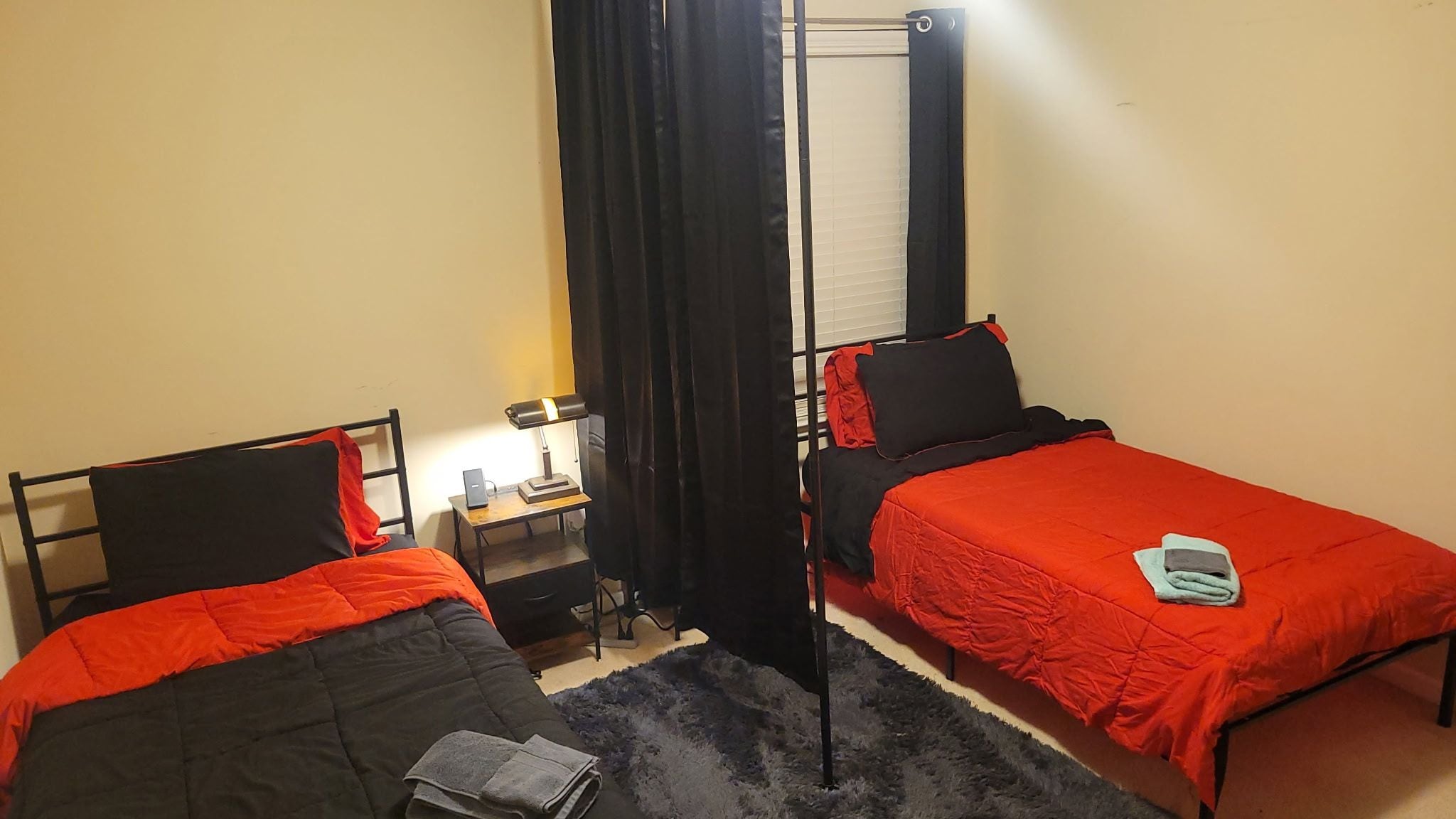 A room with two single beds, a nightstand with a lamp, a small table, and a window with curtains.