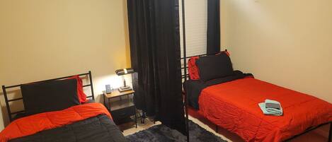 A room with two single beds, a nightstand with a lamp, a small table, and a window with curtains.