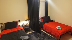 Una habitació amb dos llits individuals, una nit pota amb una làmpada, una taula petita i una finestra amb cortines.