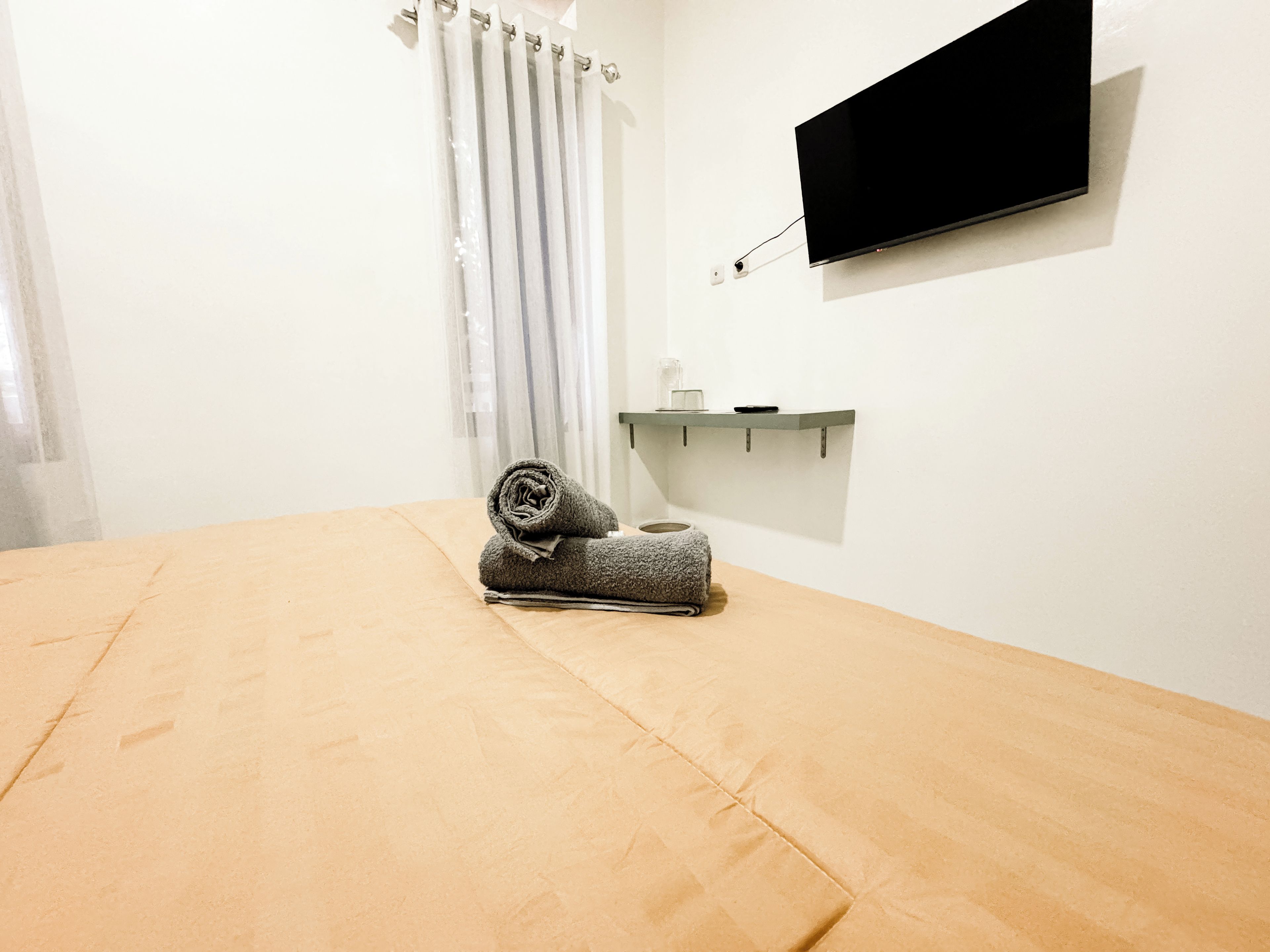 A room with a bed, a flat-screen TV mounted on the wall, a small shelf with a glass container, and a curtain.