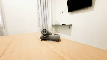 A room with a bed, a flat-screen TV mounted on the wall, a small shelf with a glass container, and a curtain.