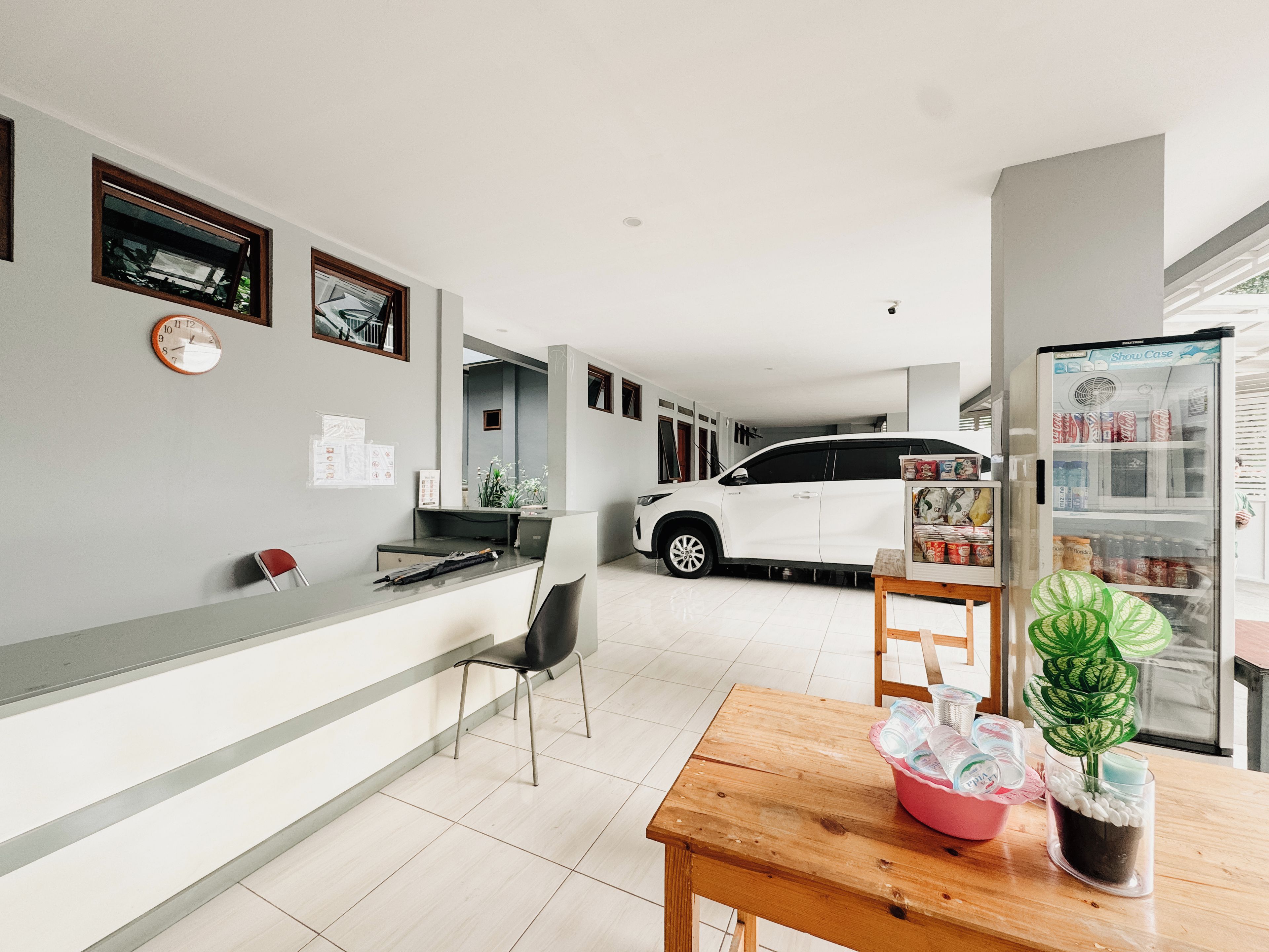 A modern interior with a reception desk, a white car parked in the background, a dining area with a wooden table, and a refrigerator stocked with beverages.