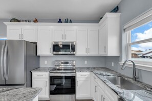 A modern kitchen with white cabinets, stainless steel appliances, a granite countertop, and a window with a view of houses. - AlohaSunset: Affordable Beach Luxury & Walk to Beach (Panama City)