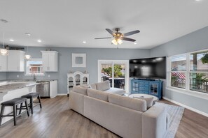 A modern kitchen with white cabinets, a dining area with a gray sofa, and a large TV. - AlohaSunset: Affordable Beach Luxury & Walk to Beach (Panama City)