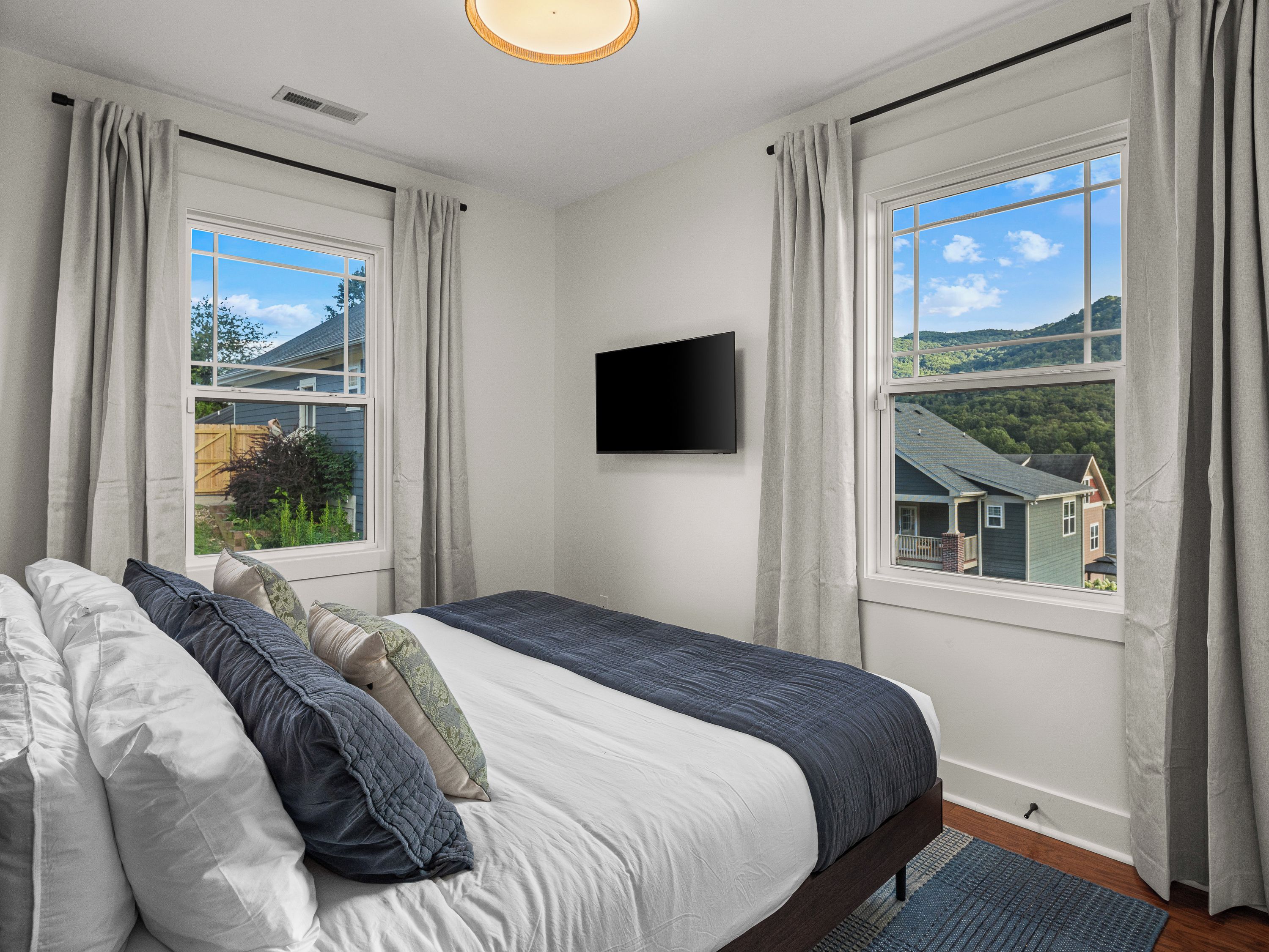 Un dormitorio con cama, cortinas, televisor de pantalla plana y vista a una casa y árboles.