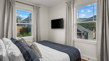 Un dormitorio con cama, cortinas, televisor de pantalla plana y vista a una casa y árboles.