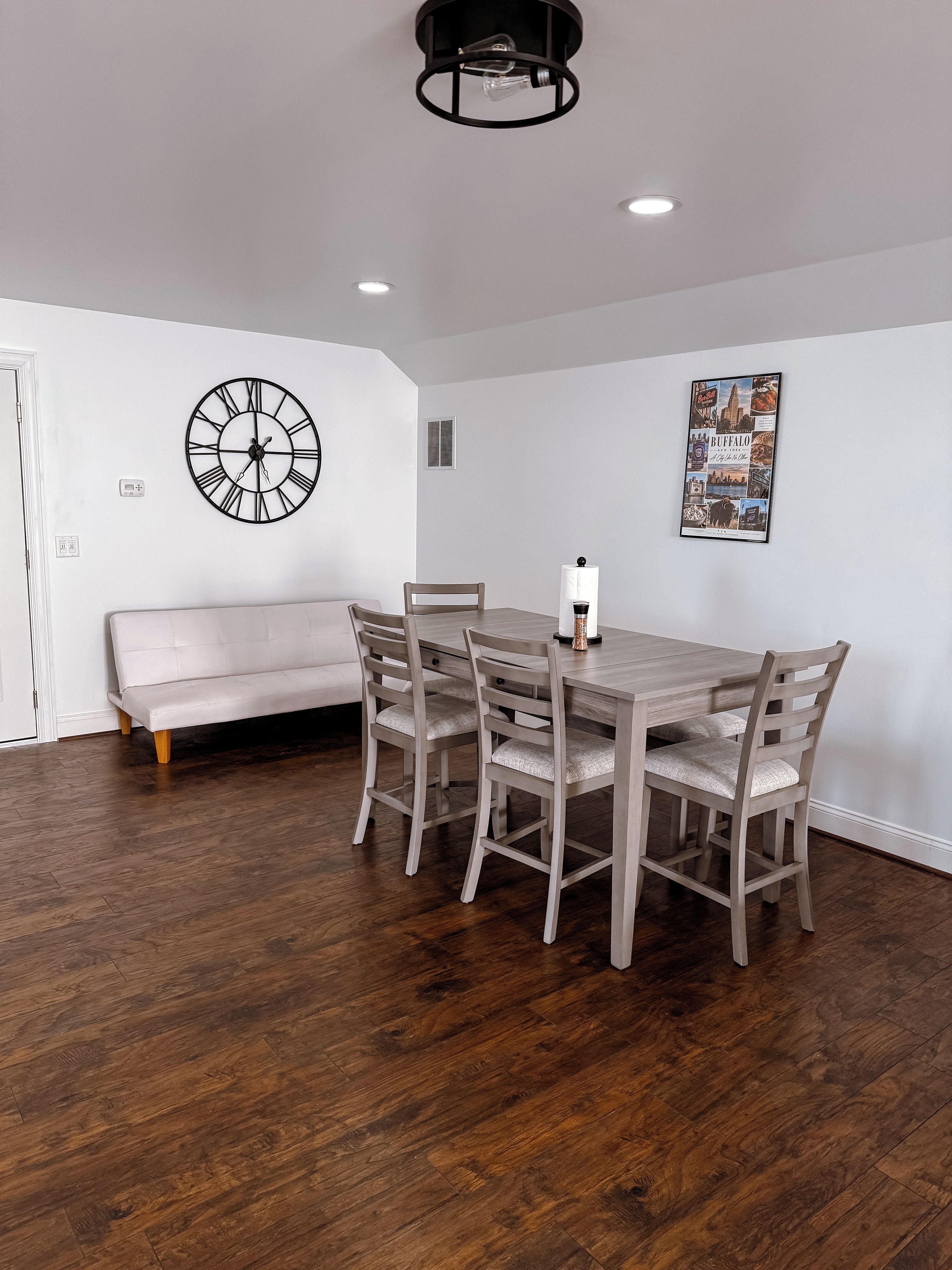 A dining area with a wooden table and chairs, a wall clock, and a framed picture.