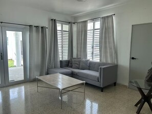 A modern living room with a grey sofa, a glass coffee table, and a sliding glass door leading to an outdoor area. - Casa Magia - 3BR Modern Home Near San Juan (Guaynabo)