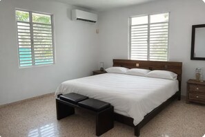 A bedroom with a bed, a bench, a nightstand, an air conditioner, and windows with blinds. - Casa Magia - 3BR Modern Home Near San Juan (Guaynabo)