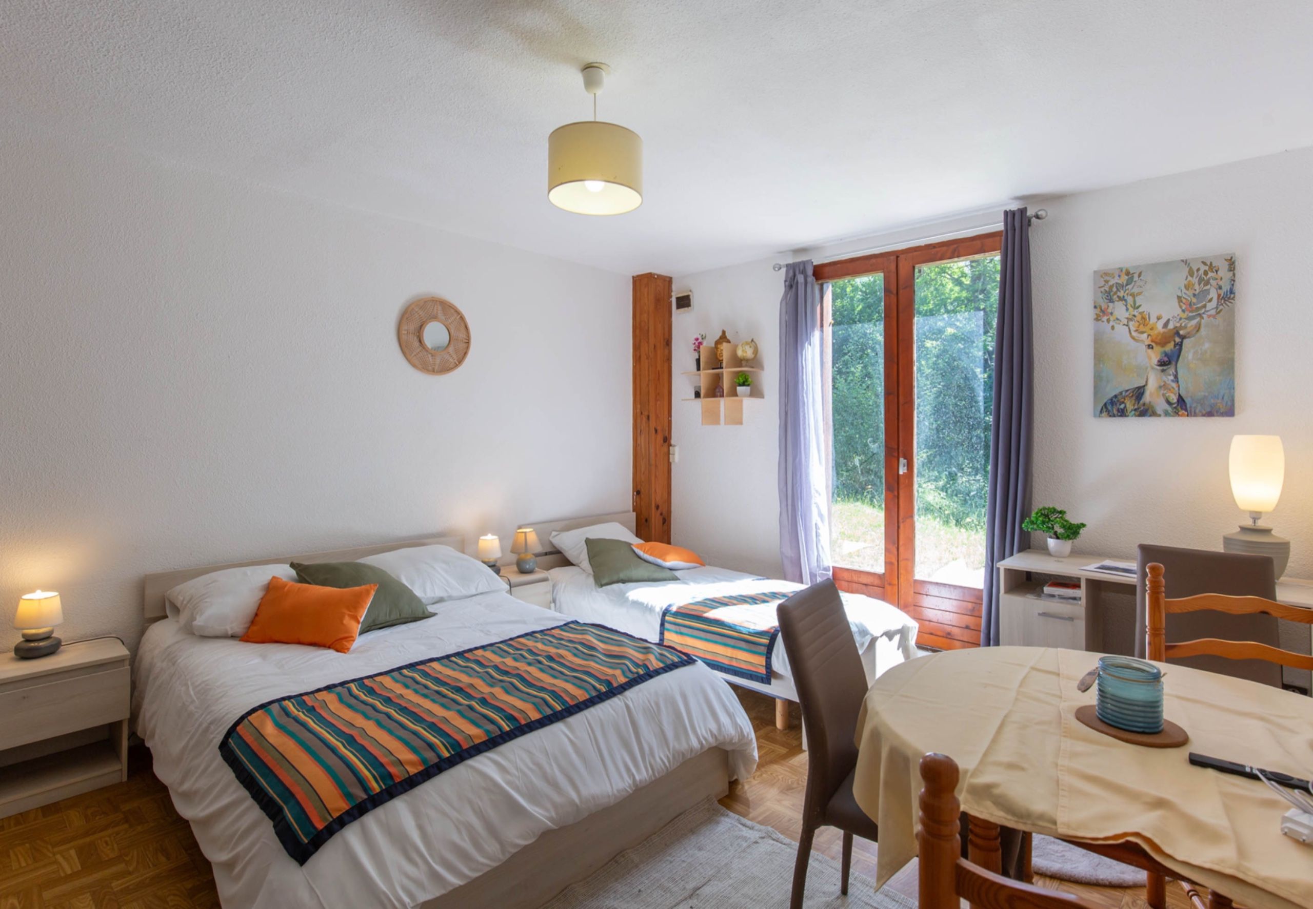 A bedroom with two beds, a dining table, a chair, a lamp, a painting, and a window with curtains.