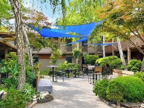 Un patio exterior con mesas y sillas, rodeado de follaje y cubierto por toldos azules.