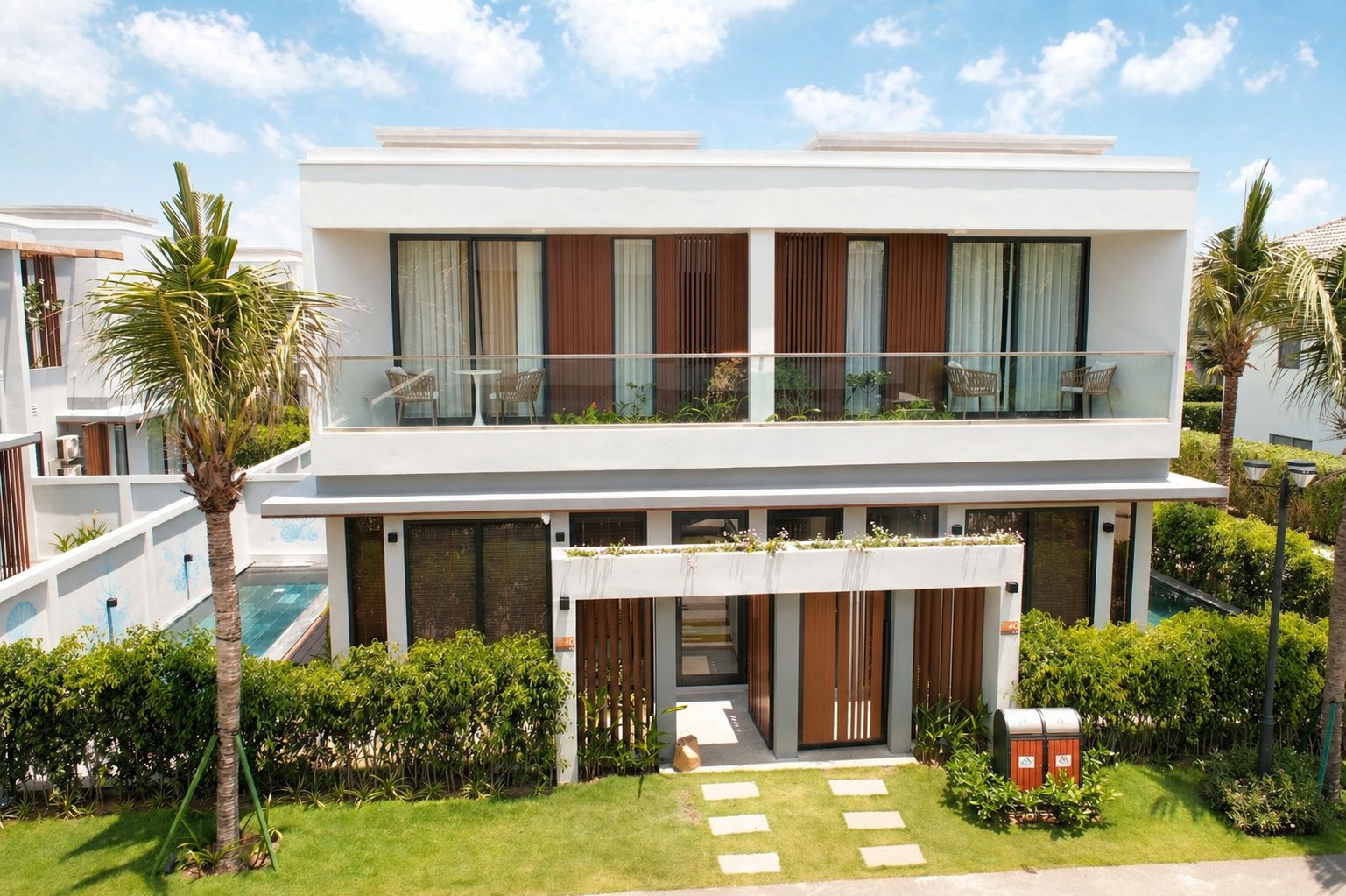 A modern two-story residential building with a balcony, a swimming pool, and lush greenery.