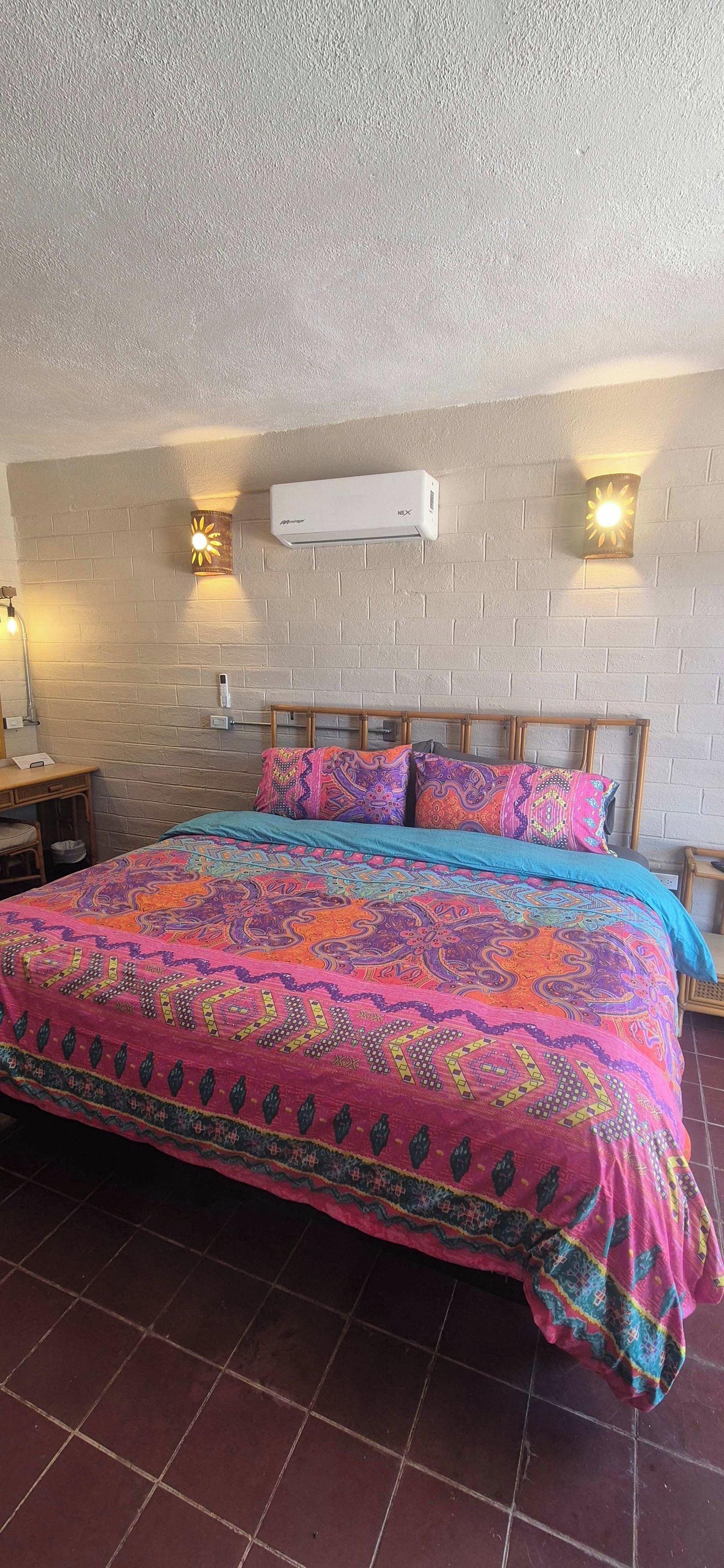 A bed with a colorful patterned bedspread, a white air conditioning unit, and two wall-mounted lamps.