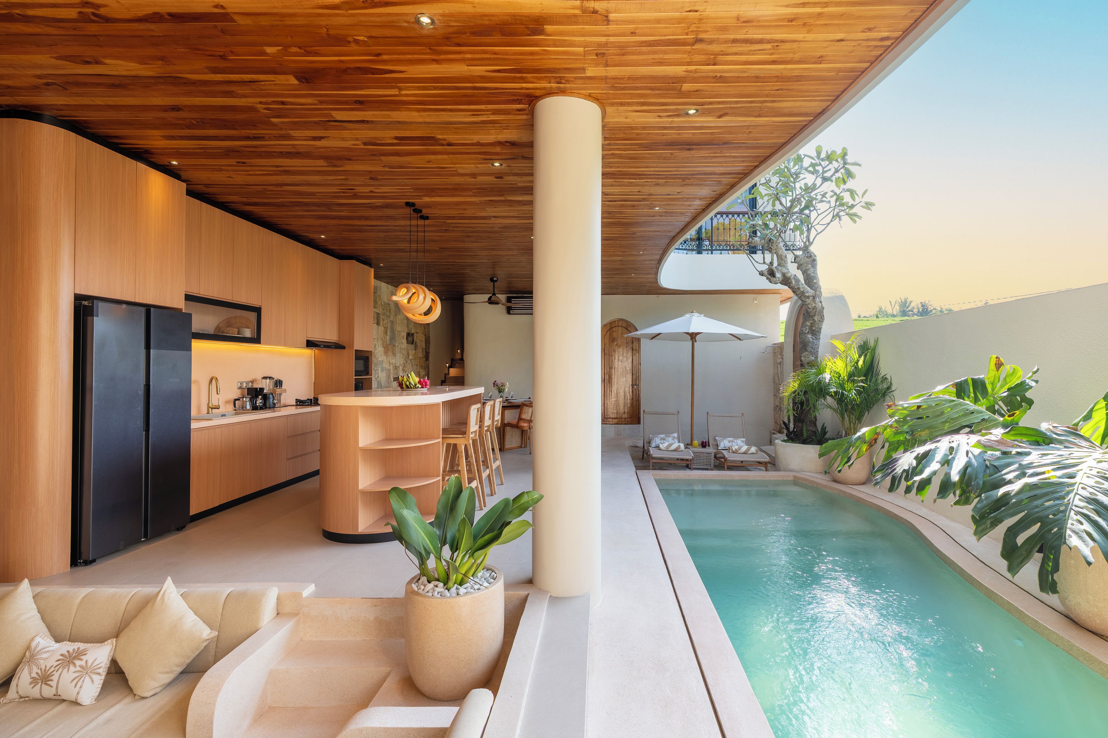 A modern kitchen with a built-in refrigerator, a wooden ceiling, and a dining area with a poolside view.