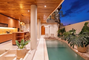Una cocina moderna con armarios de madera y un comedor con sillas y una mesa. Hay un área de piscina con sillones y un gran paraguas.