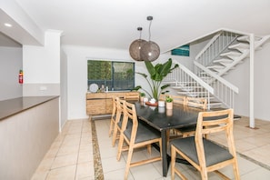A modern kitchen with a dining area, featuring a staircase and a view of greenery outside. - BREEZE AT BROADBEACH - Elite Holiday Homes (Gold Coast)