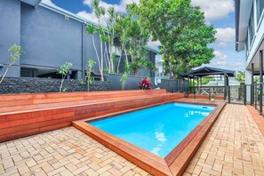 A swimming pool with a wooden deck, surrounded by a stone wall and plants. - BREEZE AT BROADBEACH - Elite Holiday Homes (Gold Coast)