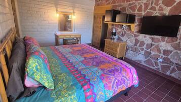 A bedroom with a bed, a wooden nightstand, a desk with a mirror, a television mounted on a stone wall, and a stone-patterned floor.