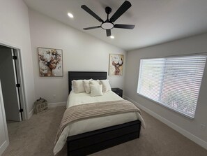 Et soveværelse med seng, ventilator, et vindue med persienner og to billeder hængt på væggen.