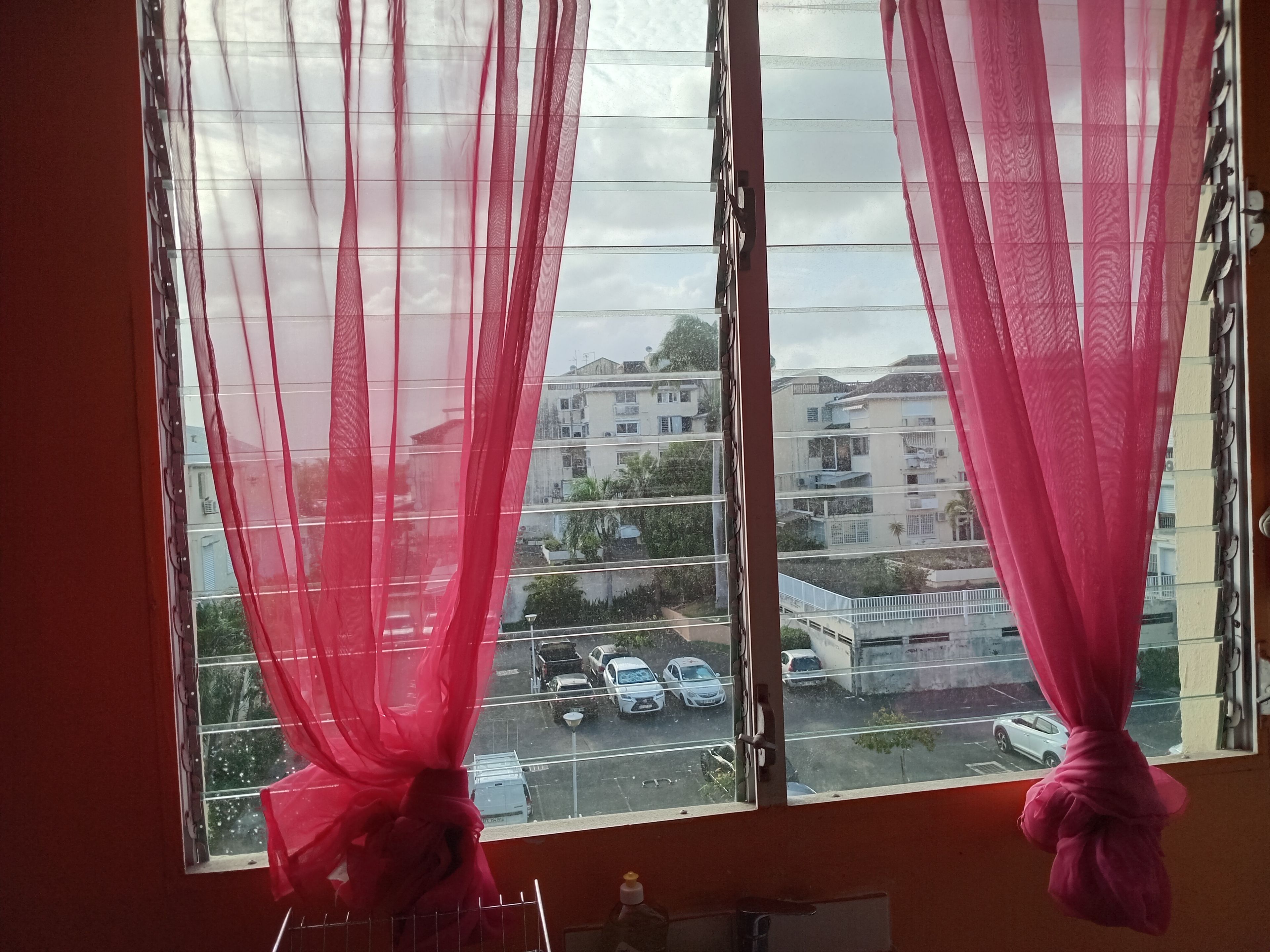 Una ventana con cortinas traslúcidas y vista a un estacionamiento y edificios.