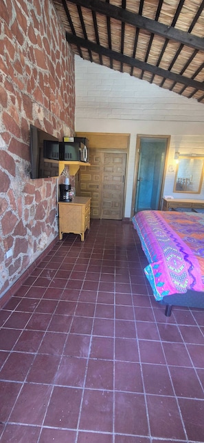 A room with a brick wall, tiled floor, a bed with a colorful blanket, a television mounted on the wall, a wooden cabinet, and a door. - Sandy Shores 9D (La Paz)