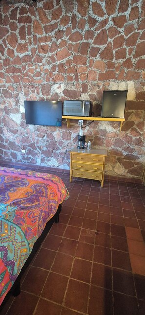 Ett rum med en vägg av sten, en säng med ett färgglatt överkast, en tv, en kaffemaskin och ett litet träbord.