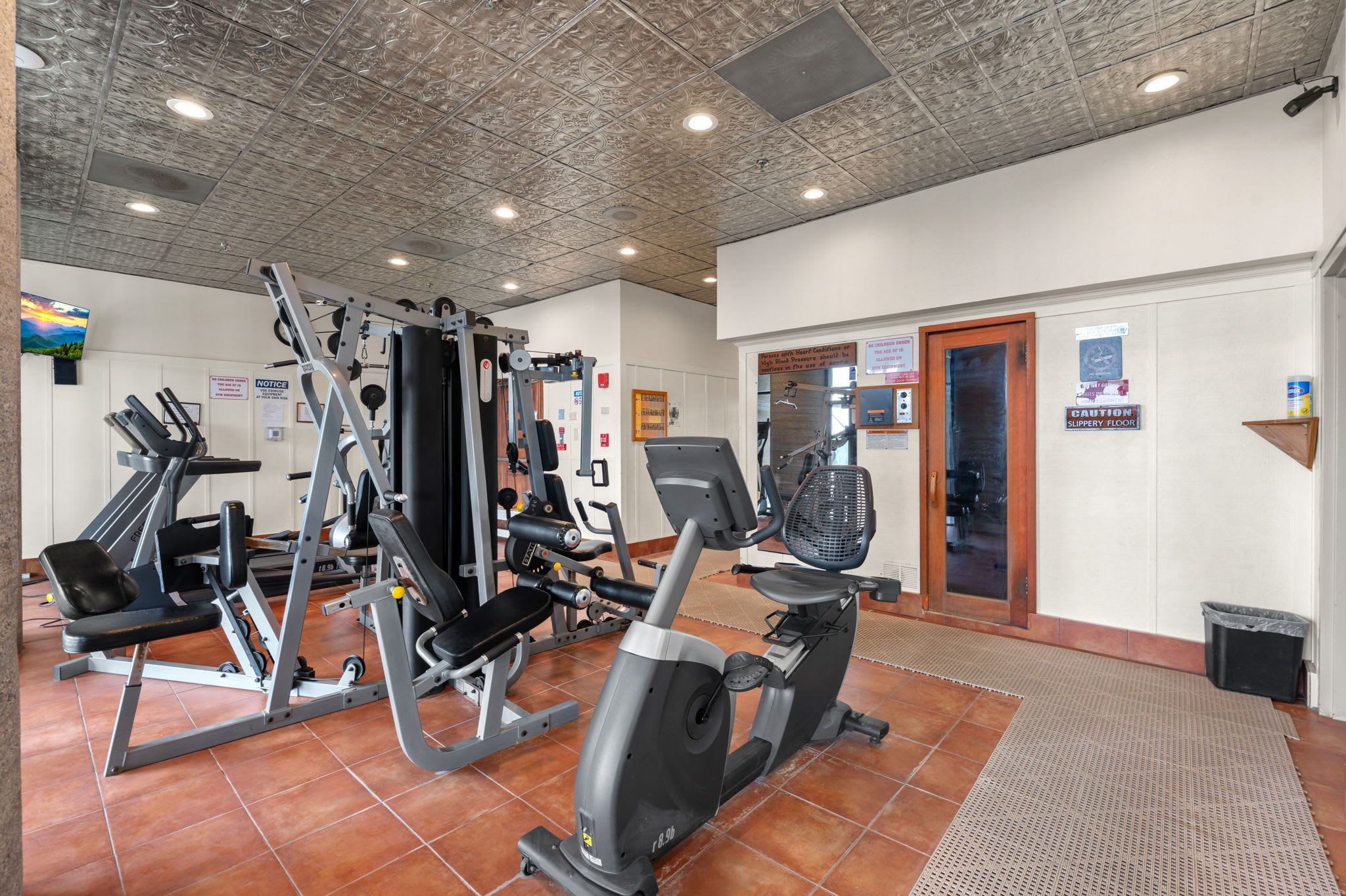 A gym with various exercise equipment, a television, and a door with a caution sign.