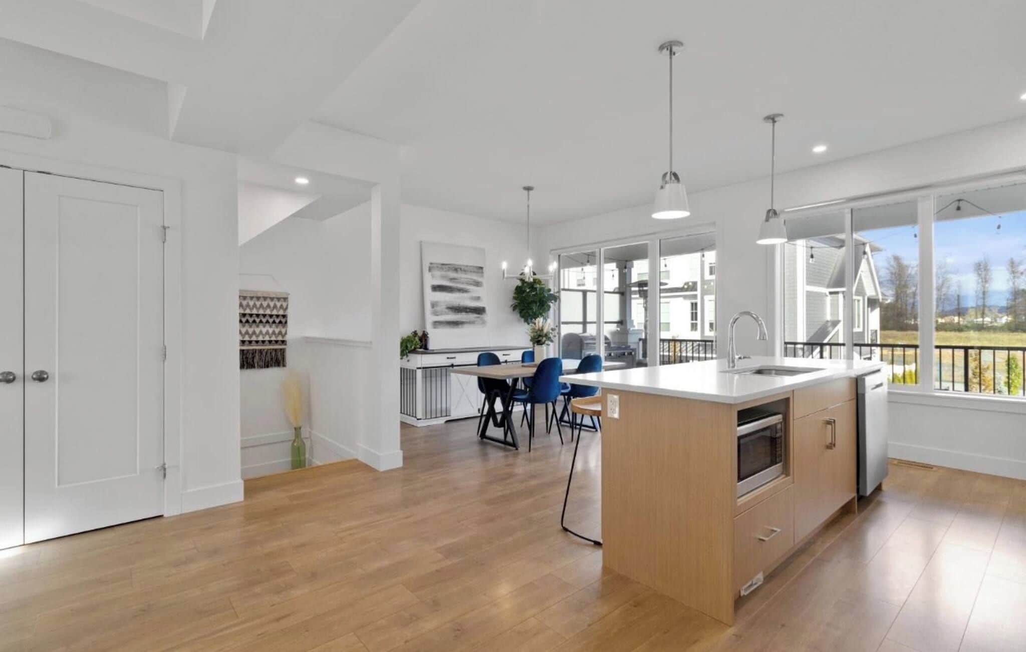 A modern kitchen with a central island, a dining area with blue chairs, and large windows offering a view of the outdoors.