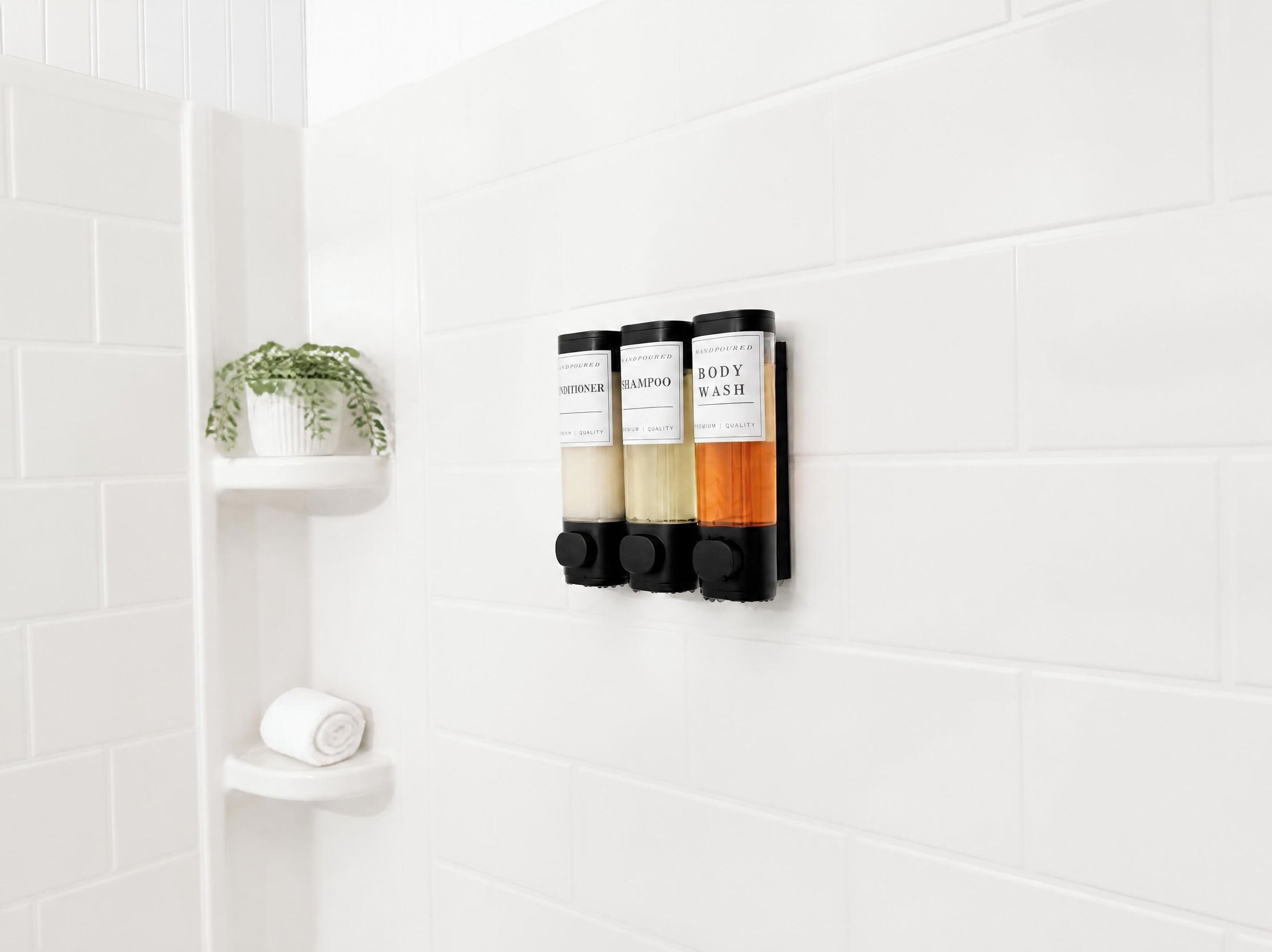 A bathroom with white tiled walls, a white shelf with a plant, and three black dispensers labeled 'HANDPOURED CONDITIONER,' 'HANDPOURED SHAMPOO,' and 'HANDPOURED BODY WASH.'