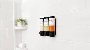 A bathroom with white tiled walls, a white shelf with a plant, and three black dispensers labeled 'HANDPOURED CONDITIONER,' 'HANDPOURED SHAMPOO,' and 'HANDPOURED BODY WASH.'