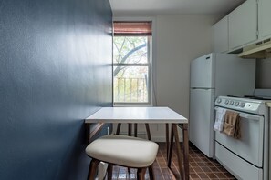 Eine kleine Küche mit Fenster, einem weißen Tisch, einem Stuhl, einem Kühlschrank und einem Backofen.