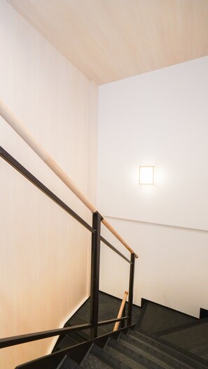 A staircase with a wooden handrail and black metal balusters, leading to a landing with a light fixture. - HANASTAY HANAZONOYA (Osaka)