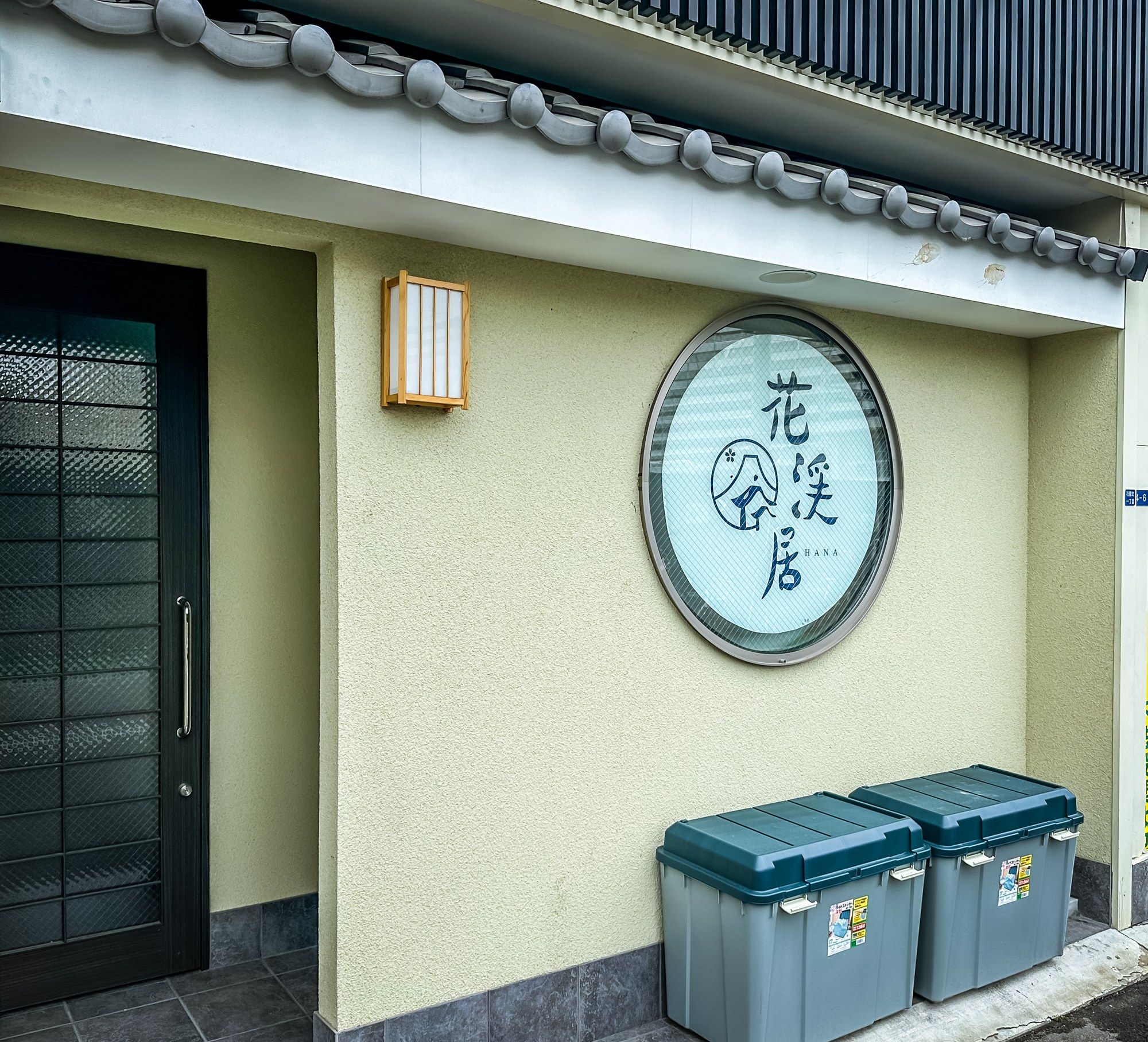 A building facade with a traditional tiled roof, a circular window with Japanese characters, and two grey storage bins.