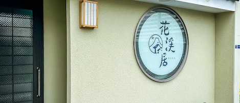 A building facade with a traditional tiled roof, a circular window with Japanese characters, and two grey storage bins.