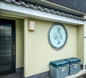 Eine Gebäudefassade mit traditioneller Ziegeldach, einem runden Fenster mit japanischen Schriftzeichen und zwei grauen Aufbewahrungsbehältern.