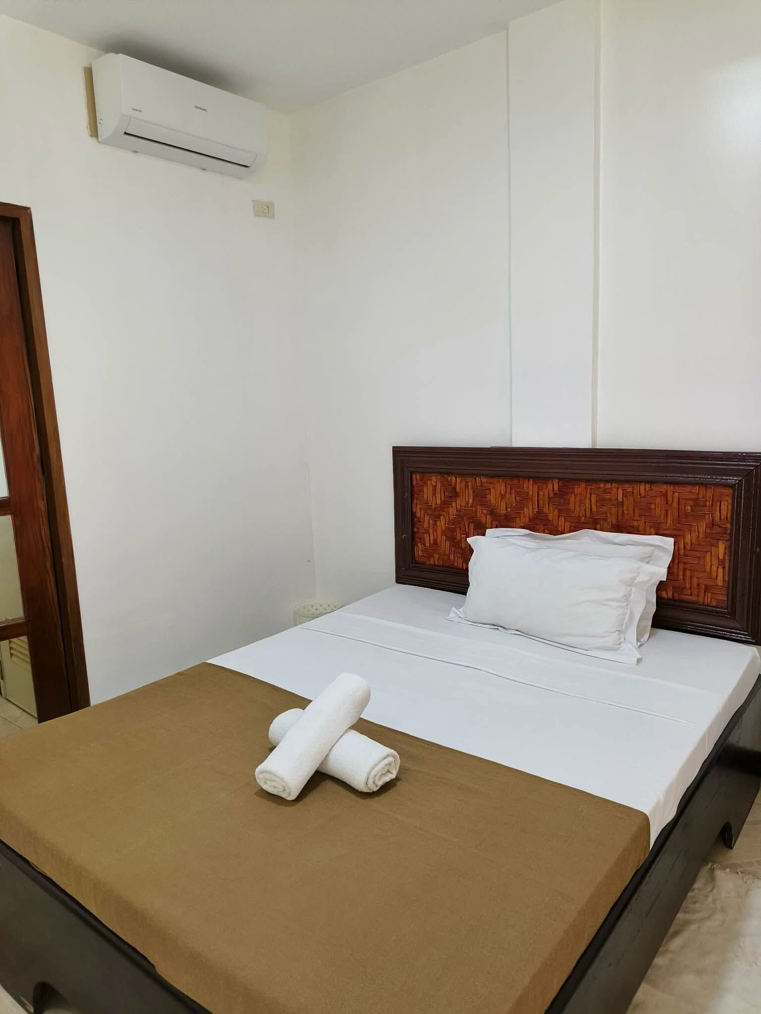 Una cama bien hecha con ropa blanca y una colcha marrón, un cabecero de madera y un equipo de aire acondicionado en la pared.