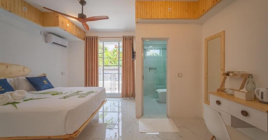 Habitación de hotel con cama, ventilador de techo, baño con puerta de vidrio y cómoda de madera.