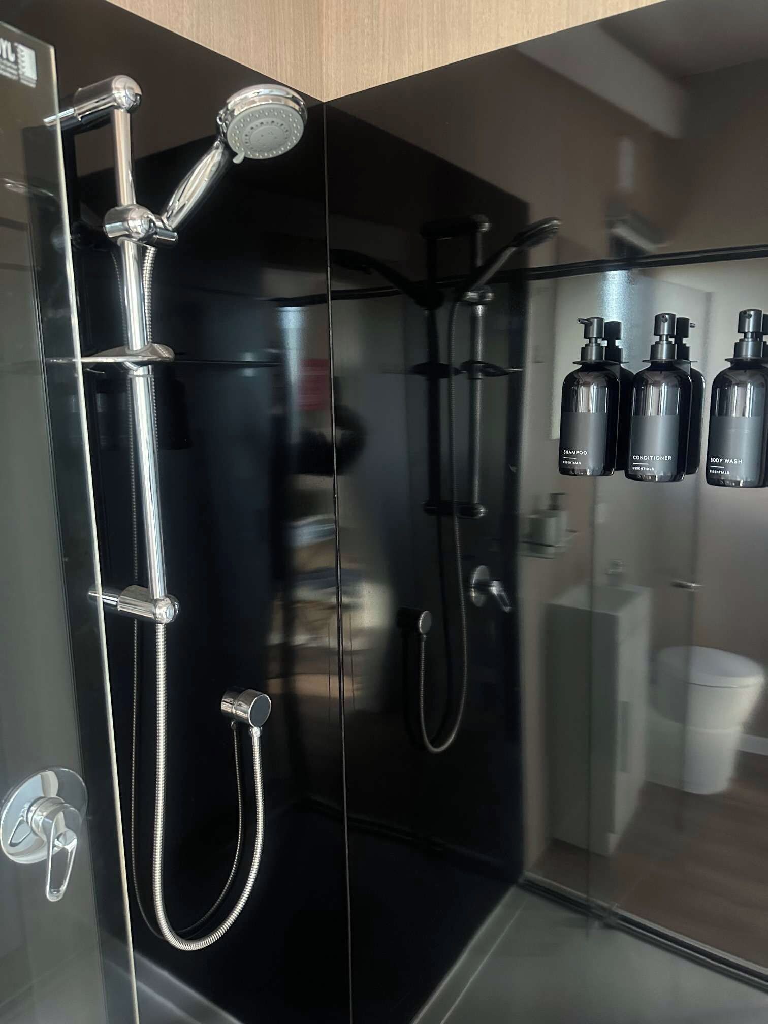 Une salle de bain moderne avec une douche à parois de verre, un pommeau de douche à main, un robinet mural et trois flacons de produits de toilette fixés au mur.